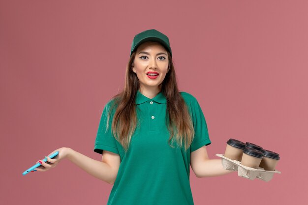 Corriere femminile di vista frontale in uniforme verde parlando al telefono e tenendo le tazze di caffè di consegna sul lavoro di consegna uniforme di servizio parete rosa