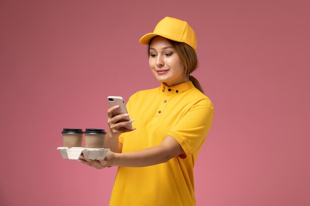 Corriere femminile di vista frontale nel capo giallo uniforme giallo che tiene le tazze di caffè usando il telefono sul lavoro di colore del lavoro di consegna uniforme del fondo rosa