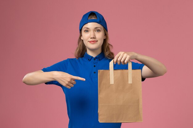Corriere femminile di vista frontale nel pacchetto di carta di consegna della tenuta del capo uniforme blu sulla parete rosa, consegna del lavoratore di servizio del dipendente