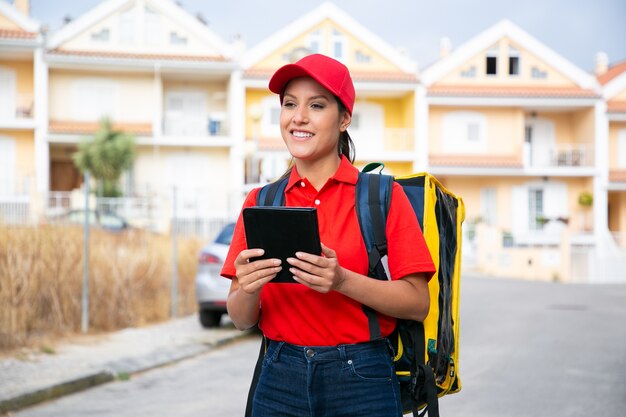 Corriere femminile felice che consegna l'ordine e che lavora nel servizio postale. Consegna sorridente in berretto rosso e camicia che trasportano zaino giallo e che tiene compressa. Servizio di consegna e concetto di acquisto online