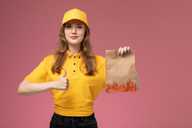 Corriere femminile giovane vista frontale in capo giallo uniforme giallo che tiene il pacchetto di consegna del cibo con il sorriso sul colore del servizio di lavoro di consegna uniforme sfondo rosa scuro