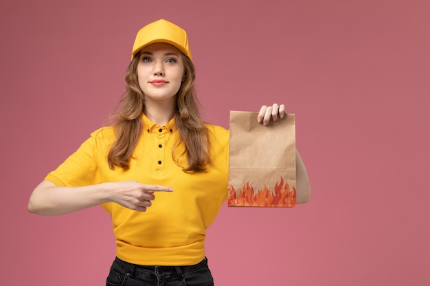 Corriere femminile giovane vista frontale in capo giallo uniforme giallo che tiene il pacchetto di consegna del cibo su colore di servizio di lavoro di consegna uniforme sfondo rosa