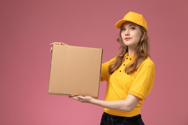 Corriere femminile giovane vista frontale in mantello giallo uniforme giallo che tiene la scatola di consegna del cibo con un leggero sorriso sul colore del servizio di lavoro di consegna uniforme da scrivania rosa scuro