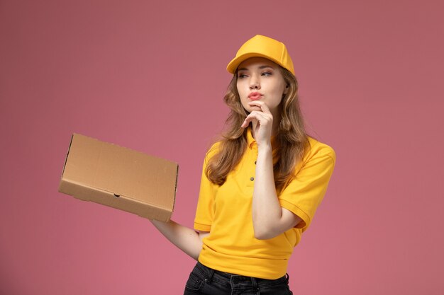 Corriere femminile giovane vista frontale in mantello giallo uniforme giallo che tiene la scatola di consegna del cibo sul colore di servizio di lavoro di consegna uniforme sfondo rosa scuro