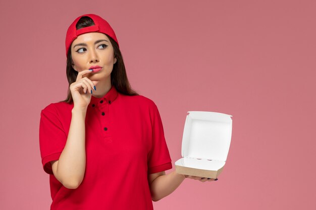 Corriere femminile giovane vista frontale in uniforme rossa e mantello con poco pacchetto di cibo di consegna sulle sue mani pensando sulla parete rosa chiaro