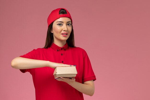 Corriere femminile giovane vista frontale in uniforme rossa e mantello con poco pacchetto di cibo di consegna sulle sue mani sul muro rosa