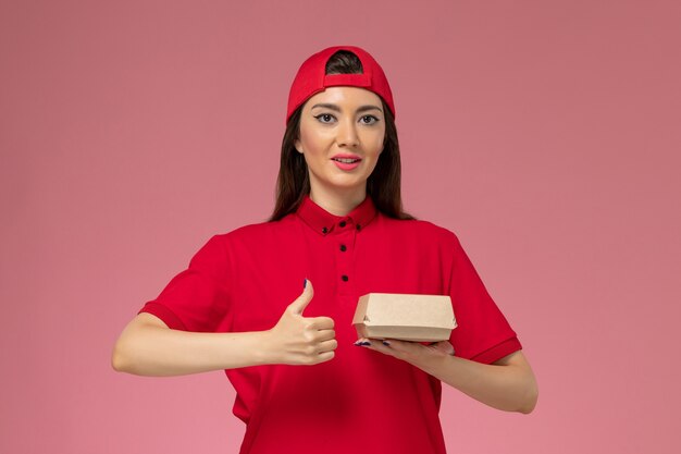 Corriere femminile giovane vista frontale in uniforme rossa e mantello con poco pacchetto di cibo di consegna sulle sue mani sul muro rosa