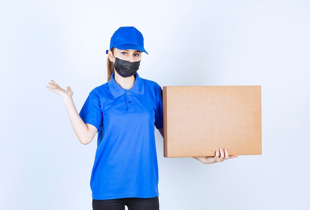 Corriere femminile in maschera e uniforme blu che tiene un grosso pacco di cartone e indica qualcuno