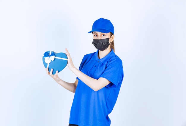 Corriere femminile in maschera e uniforme blu che tiene una confezione regalo a forma di cuore.
