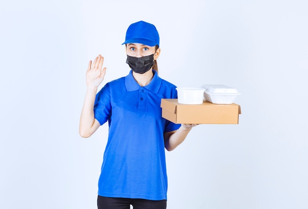 Corriere femminile in maschera e uniforme blu che tiene una scatola di cartone e pacchi da asporto e ferma qualcuno.