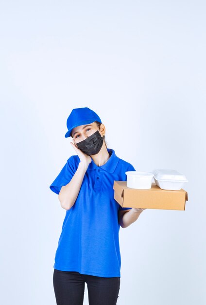 Corriere femminile in maschera e uniforme blu che tiene una scatola di cartone e pacchi da asporto e sembra assonnato e stanco