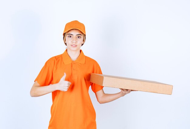 Corriere femminile in uniforme arancione che consegna una scatola di cartone e mostra il segno della mano di soddisfazione