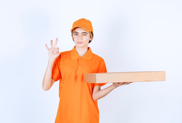 Corriere femminile in uniforme arancione che consegna una scatola di cartone e mostra il segno della mano di soddisfazione