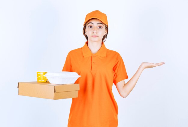 Corriere femminile in uniforme arancione con scatole da asporto gialle e bianche con un pacco di cartone e sembra confuso e pensieroso