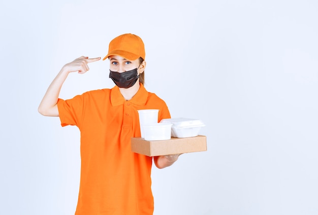 Corriere femminile in uniforme arancione e maschera nera con in mano un pacco di cartone, cibo e bevande da asporto mentre pensa e pianifica