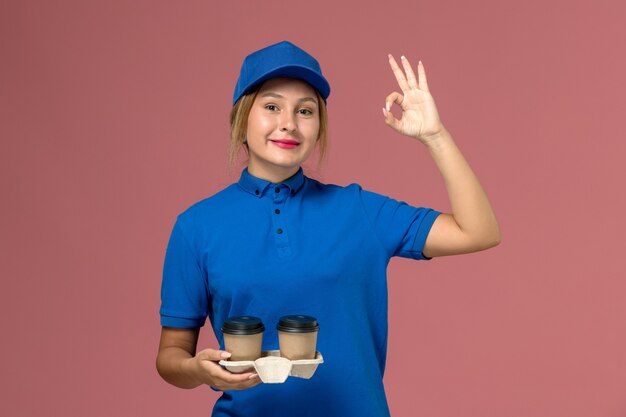 corriere femminile in uniforme blu che posa e che tiene le tazze di caffè con un sorriso sul rosa, lavoratore di consegna uniforme di servizio