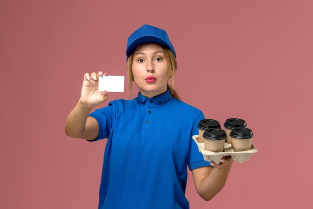 corriere femminile in uniforme blu che tiene carta bianca insieme a tazze di caffè marrone su rosa chiaro, consegna uniforme di lavoro di servizio