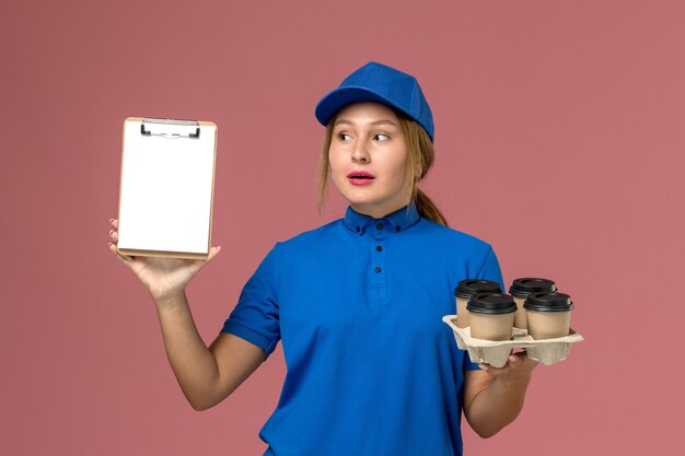 corriere femminile in uniforme blu che tiene il blocco note insieme a tazze di caffè marrone su rosa chiaro, consegna uniforme di servizio