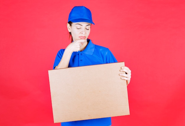 Corriere femminile in uniforme blu che tiene in mano una scatola di cartone per pizza da asporto e pensa o sembra confuso.