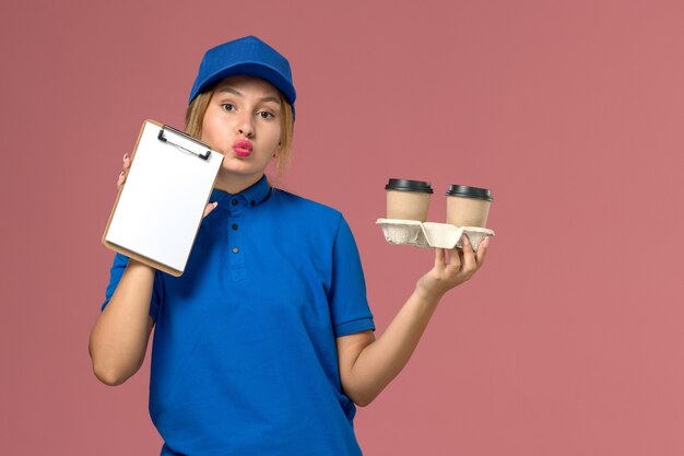 corriere femminile in uniforme blu che tiene le tazze di consegna di caffè e blocco note pensando al lavoro di consegna uniforme rosa, lavoratore di servizio
