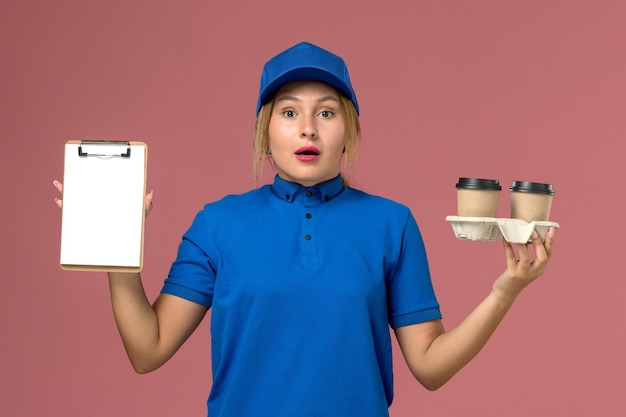 corriere femminile in uniforme blu che tiene le tazze di consegna di caffè e blocco note sul rosa, consegna uniforme dei lavoratori di servizio