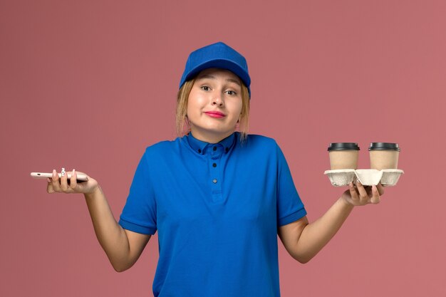 corriere femminile in uniforme blu che tiene le tazze di consegna di caffè e il suo telefono in rosa, consegna uniforme di lavoro del lavoratore di servizio