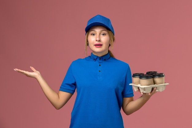 corriere femminile in uniforme blu che tiene le tazze di consegna di caffè in posa sul rosa, consegna uniforme dei lavoratori dei servizi