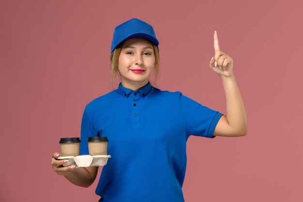 corriere femminile in uniforme blu che tiene le tazze di consegna di caffè sorridente con il dito alzato sul lavoro di consegna rosa, uniforme di servizio