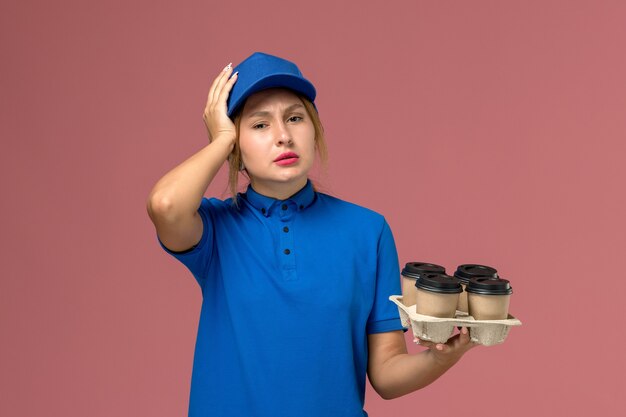 corriere femminile in uniforme blu che tiene le tazze di consegna marroni di caffè con mal di testa sulla consegna rosa, uniforme dei lavoratori di servizio