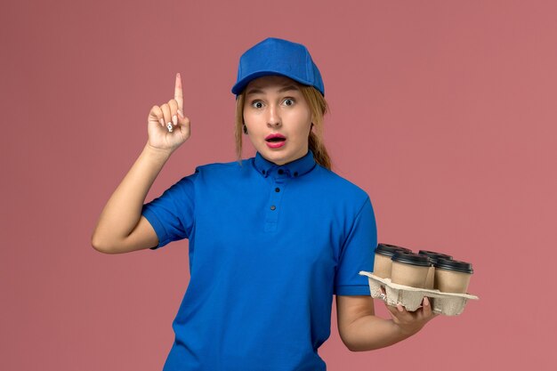 corriere femminile in uniforme blu che tiene le tazze di consegna marroni di caffè in posa con il dito alzato sul rosa, consegna uniforme di servizio