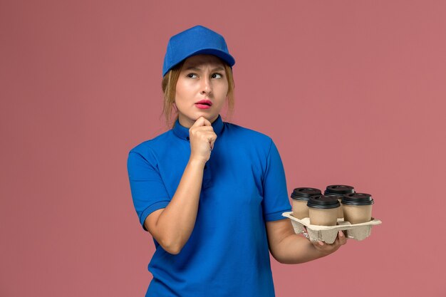 corriere femminile in uniforme blu che tiene le tazze di consegna marroni di caffè pensando in rosa, lavoratore di lavoro di consegna uniforme di servizio