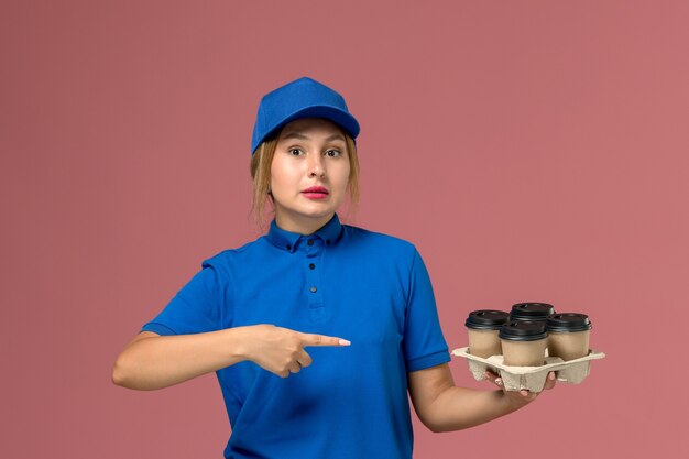 corriere femminile in uniforme blu che tiene le tazze marroni di consegna di caffè sottolineando il rosa, lavoratore di lavoro di consegna uniforme di servizio