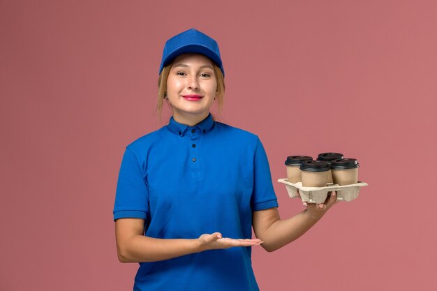 corriere femminile in uniforme blu che tiene tazze di caffè marrone consegna con un leggero sorriso sul lavoro di consegna rosa, uniforme di servizio