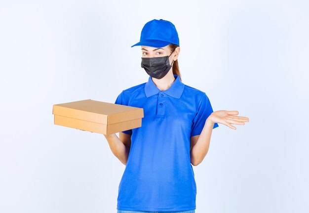 Corriere femminile in uniforme blu e maschera facciale che tiene una scatola di cartone e indica da qualche parte.