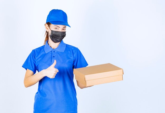 Corriere femminile in uniforme blu e maschera facciale che tiene una scatola di cartone e mostra il segno della mano di godimento.