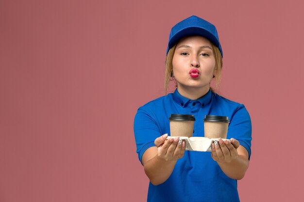 corriere femminile in uniforme blu in posa e che tiene tazze di caffè baciare sul rosa, lavoro di consegna uniforme di servizio