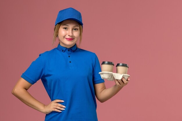 corriere femminile in uniforme blu in posa tenendo tazze di caffè con un leggero sorriso sul rosa, lavoratore di lavoro di consegna uniforme di servizio