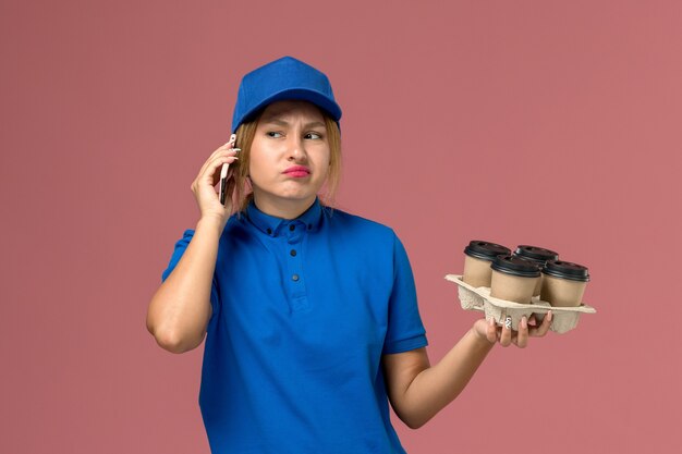 corriere femminile in uniforme blu parlando al telefono e tenendo le tazze di caffè marrone su rosa chiaro, consegna uniforme di servizio di lavoro