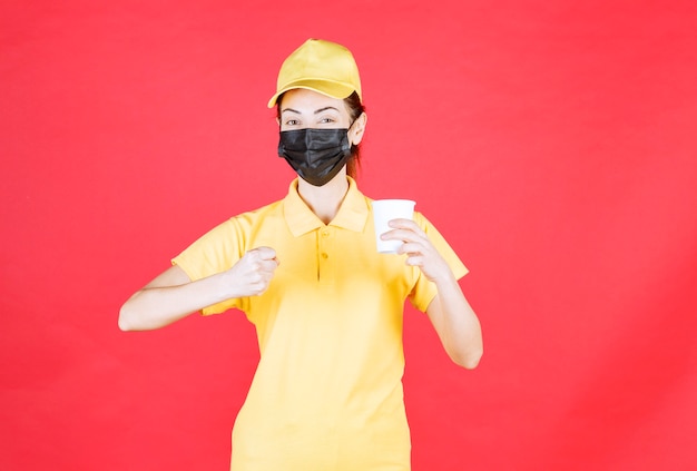 Corriere femminile in uniforme gialla e maschera nera che tiene una tazza da asporto e mostra il pugno