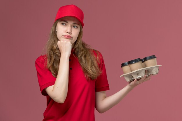 corriere femminile in uniforme rossa che pensa che tiene le tazze di caffè sul rosa, lavoratore di lavoro di consegna di servizio uniforme