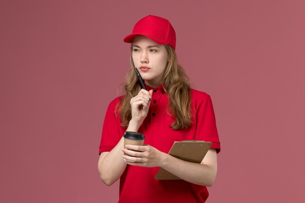 corriere femminile in uniforme rossa che tiene il blocco note e la tazza di caffè con l'espressione di pensiero sul lavoro rosa, uniforme dell'operaio di consegna del servizio