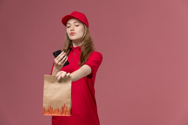 corriere femminile in uniforme rossa che tiene il pacchetto di cibo e utilizza il suo telefono su rosa chiaro, consegna del lavoratore di servizio uniforme di lavoro