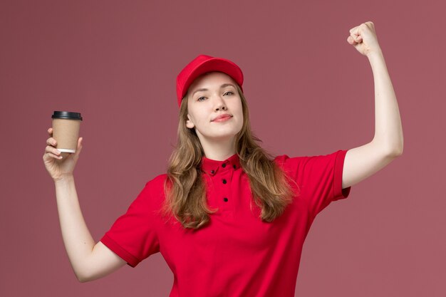 corriere femminile in uniforme rossa che tiene la tazza di caffè e che flette sul rosa chiaro, consegna dei lavoratori di servizio dell'uniforme di lavoro