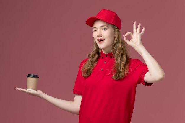 corriere femminile in uniforme rossa che tiene la tazza di caffè marrone di consegna sulla consegna dei lavoratori di servizio di colore rosa chiaro, uniforme di lavoro