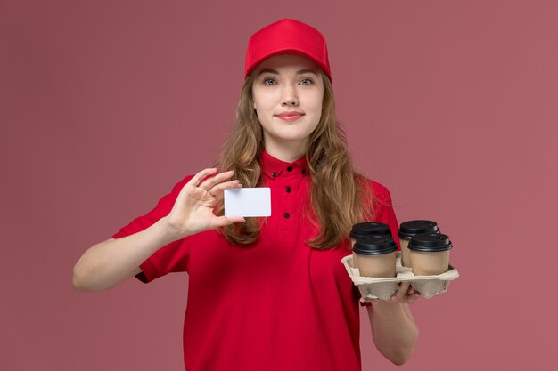 corriere femminile in uniforme rossa che tiene le tazze di caffè di consegna e carta di plastica su rosa chiaro, ragazza di consegna del lavoratore di servizio di lavoro uniforme