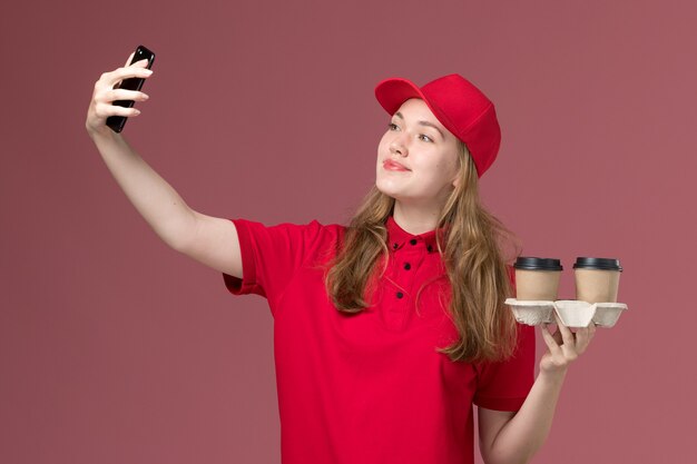 corriere femminile in uniforme rossa che tiene le tazze di caffè di consegna marroni che prendono un selfie sul lavoratore rosa, uniforme di consegna del servizio