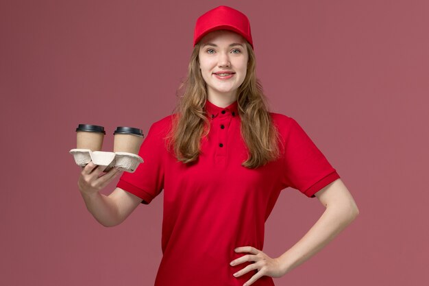 corriere femminile in uniforme rossa che tiene le tazze di caffè marroni di consegna sul lavoratore di servizio di lavoro rosa e uniforme