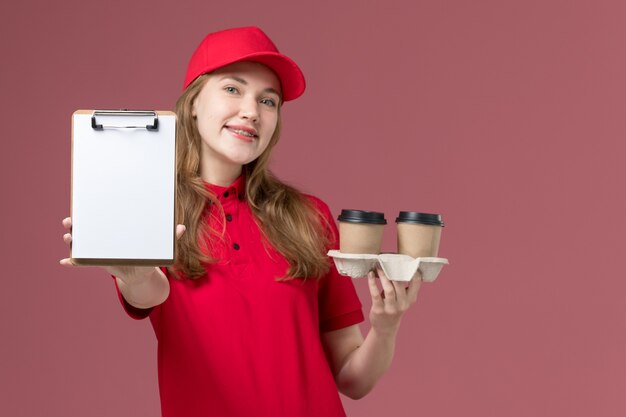 corriere femminile in uniforme rossa che tiene tazze di caffè e blocco note con il sorriso sul lavoro di consegna del servizio rosa, uniforme