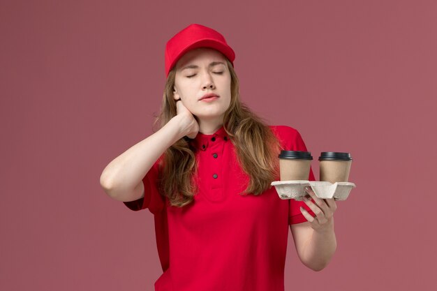 corriere femminile in uniforme rossa che tiene tazze di caffè marroni che hanno mal di collo sul lavoro dell'operaio di consegna di servizio rosa e uniforme