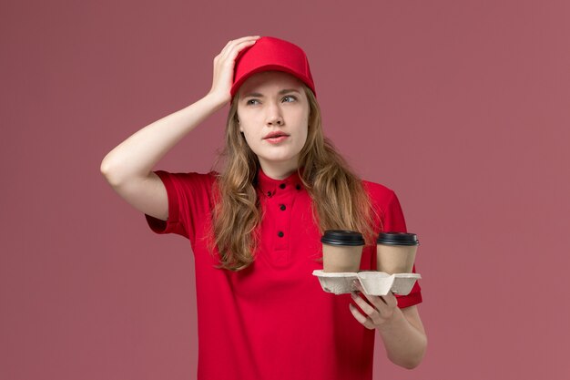 corriere femminile in uniforme rossa che tiene tazze di caffè pensando in profondità sul lavoro di consegna del servizio rosa e uniforme